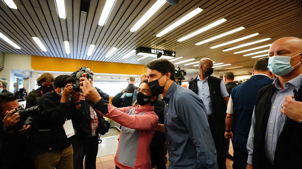 M. Trudeau, de profil, se fait prendre en photo (égoportrait) aux côtés d'une femme. Les deux sourient et portent le masque. Ils sont entourés de gardes de sécurité et de journalistes.