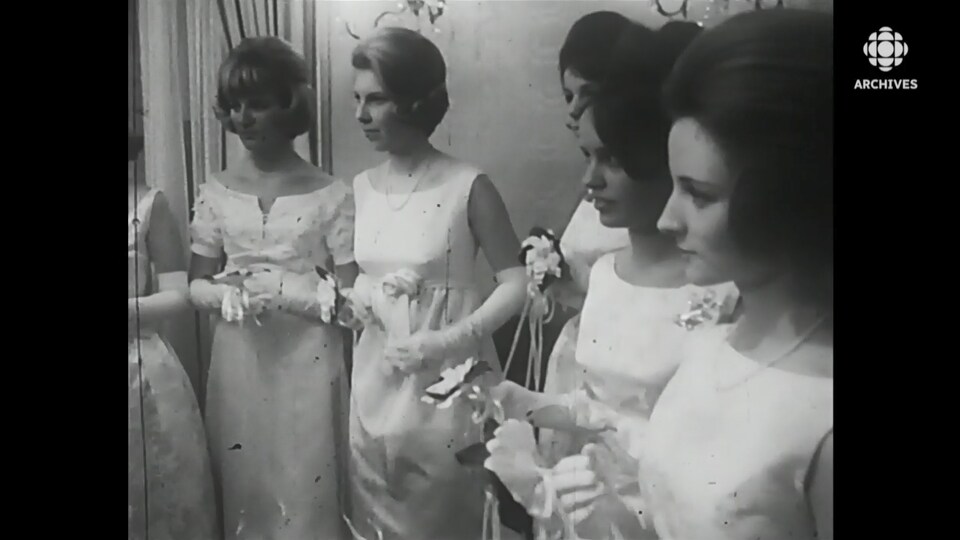 Une rangée de jeunes filles gantées et habillées de robes longues écoutent des consignes.