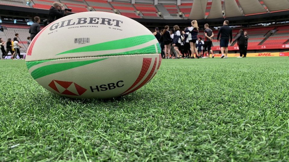 Un ballon de rugby en avant-plan sur le gazon artificiel du terrain du stade BC Place. Un groupe de joueurs de rugby en arrière-plan.