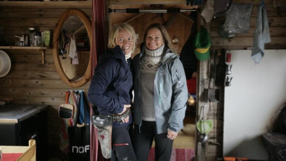Deux femmes debout dans une habitation en bois. Elle regarde la caméra.