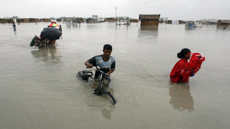 Une inondation détruit un village. Un homme pousse son vélo dans l’eau jusqu’à la taille. Une femme transporte ses biens.