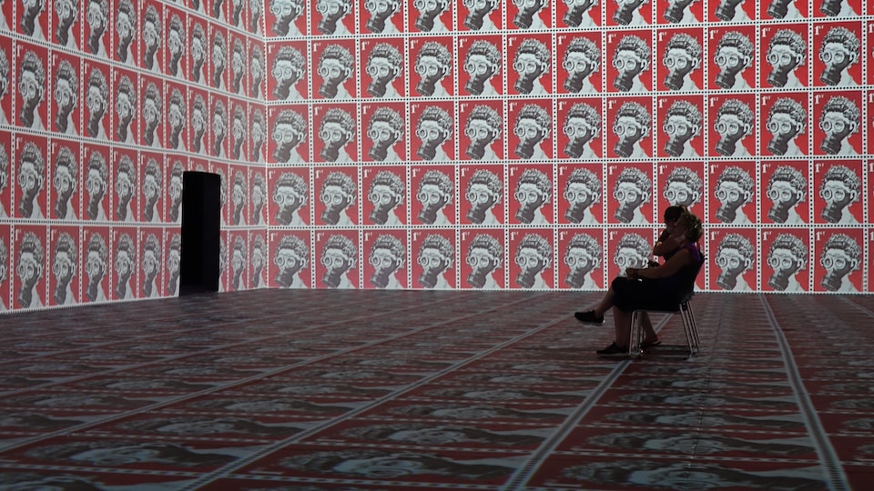 Deux personnes sont assises dans une pièce dont les murs et le plancher sont couvertes d'images de la reine d'Angleterre portant un masque à gaz.