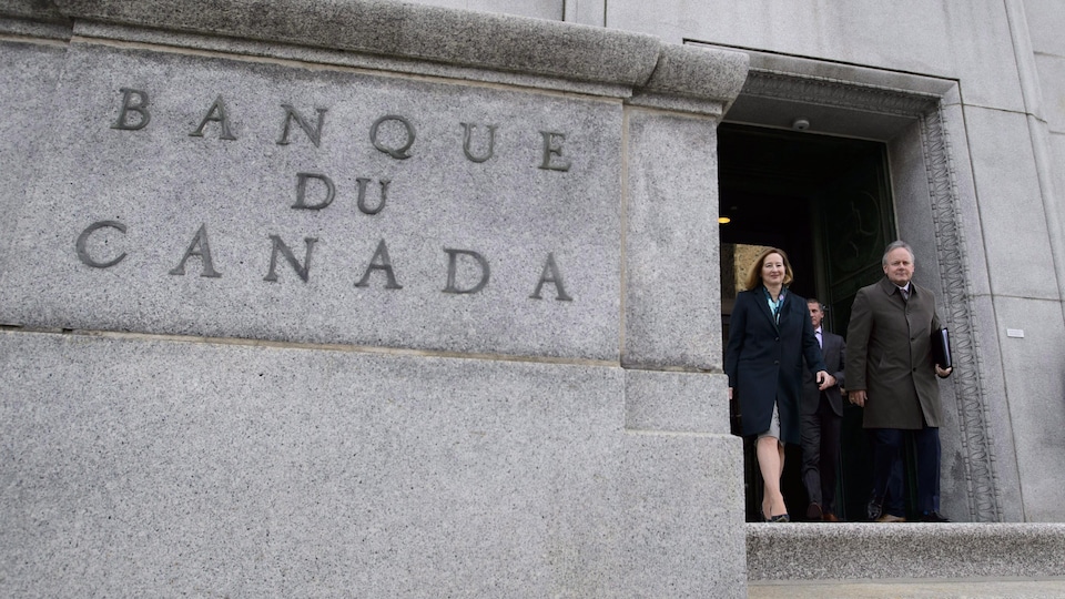 Stephen Poloz, gouverneur de la Banque du Canada et Carolyn Wilkins, première sous-gouverneure, sortent en souriant d'un immeuble sur lequel il est inscrit « Banque du Canada ».