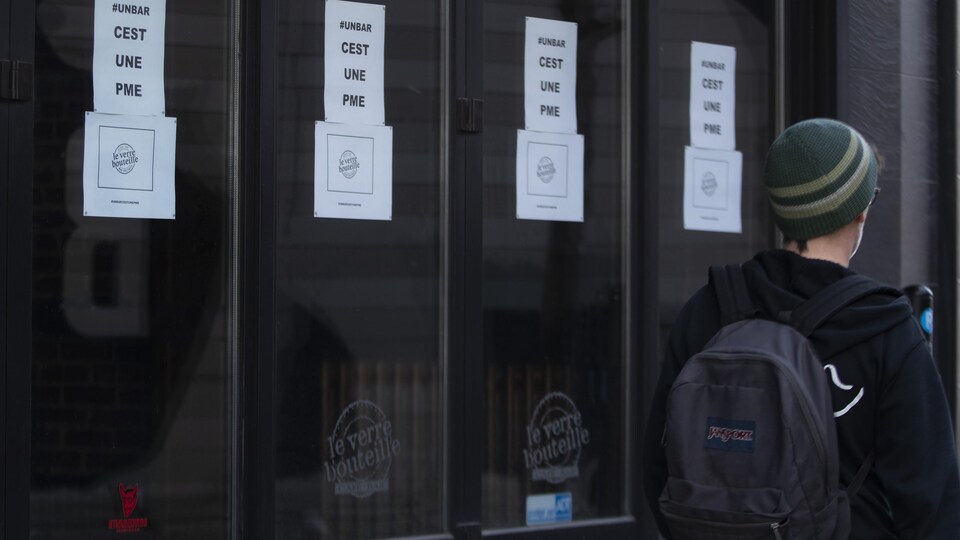 Un homme passe devant la vitrine d'un commerce où il y a une affiche sur laquelle est écrit : #unbar c'est une pme.