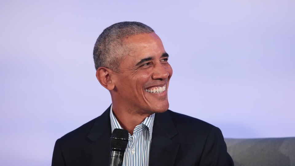 Barack Obama, micro à la main, sourit à quelqu'un à sa gauche.
