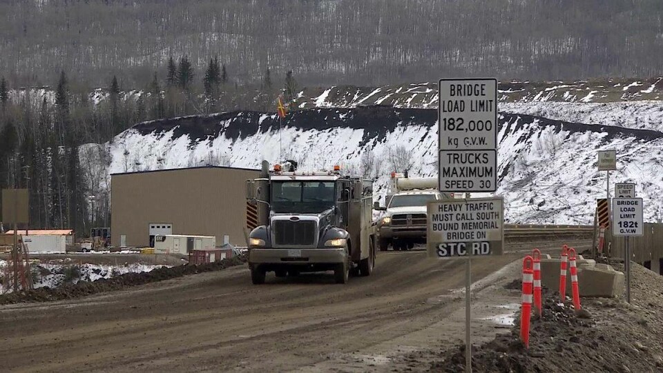 Une pancarte routière, avec des camions sur une route, en arrière-plan, un bâtiment et une colline avec des arbres et de la neige.