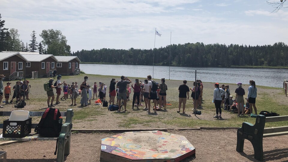 Près des chalets et du lac, plusieurs personnes écoutent des consignes avant une activité.