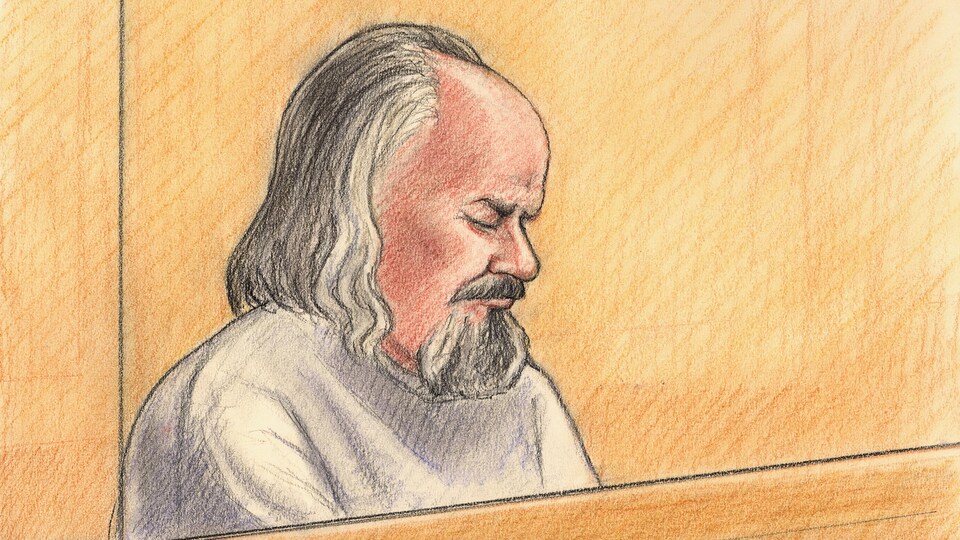 Un homme au cheveux longs et gris est assis dans le box des accusés, les yeux fermés. Il porte également une longue barbe.