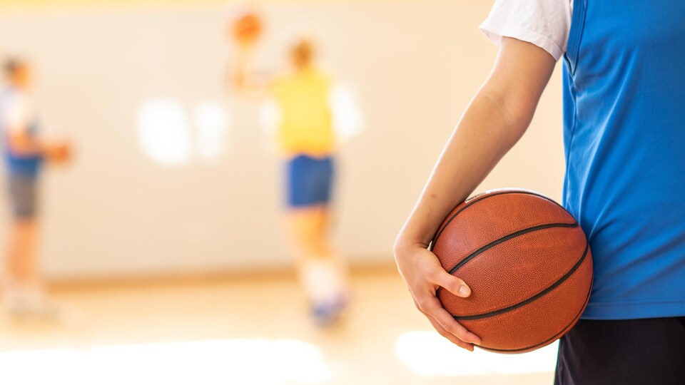 Une femme, anonyme, tient un ballon de basketball. Elle est dans un gymnase et porte un chandail de pratique de couleur bleu.