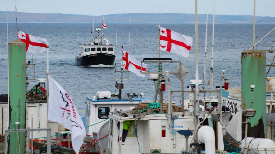 Un bateau de pêche au large, devant des bateaux avec des drapeaux micmacs au quai.