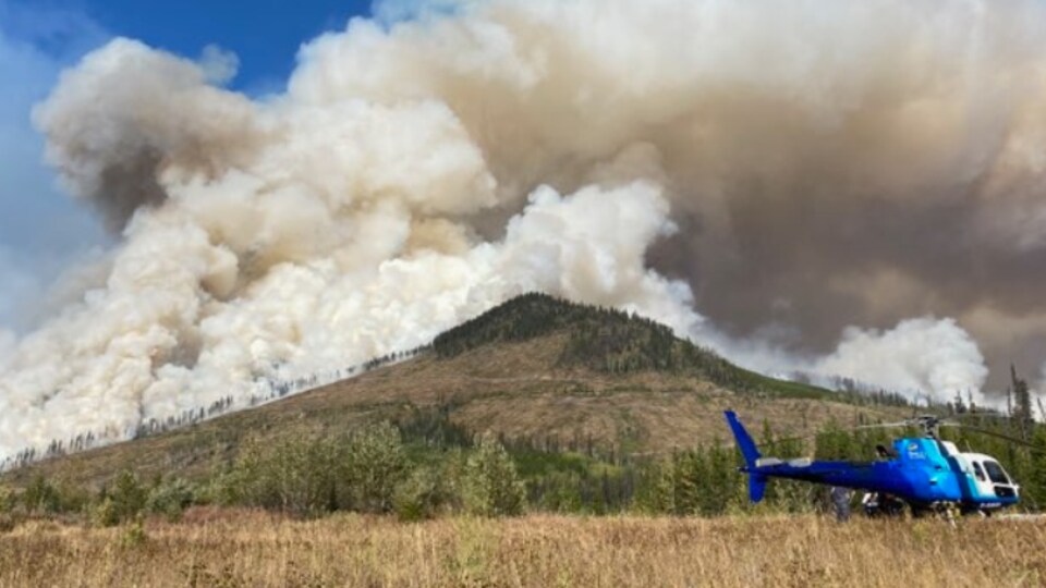 Des panaches de fumée du feu de forêt Battleship Mountain derrière une montagne et un hélicoptère qui est posé sur un champs.