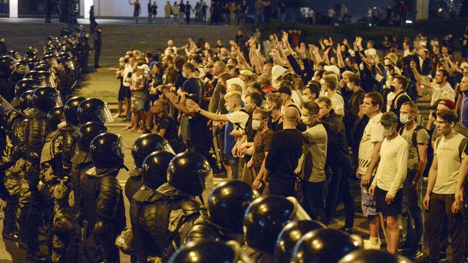 Des civils, masqués pour la plupart, font face à des policiers antiémeutes disposés en rangé dans une rue. 