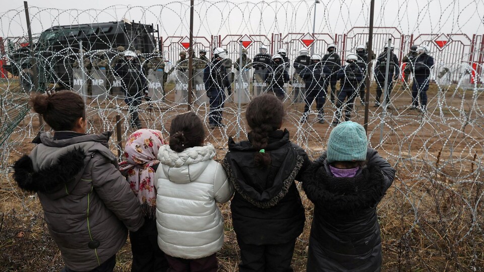 Des enfants sont derrière des barbelés et des militaires les surveillent de l'autre côté du fil métallique.