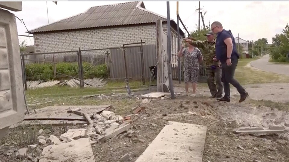 Deux civils et un homme en uniforme devant une résidence endommagée lors d'affrontements armés.
