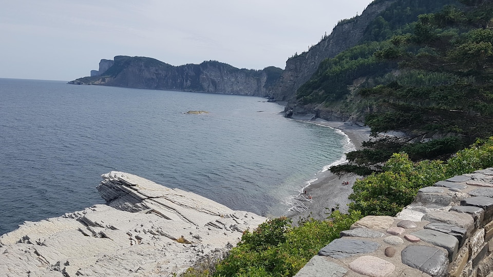 Muret de pierre devant une plage de galets, au pied d'une forêt à flanc de montagne.