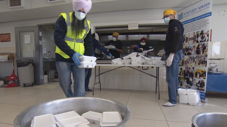 Plusieurs bénévoles, portant des turbans, des masques et des gants, dans une cuisine communautaire, mettent de la nourriture dans des boites blanches et les placent dans un grand conteneur.