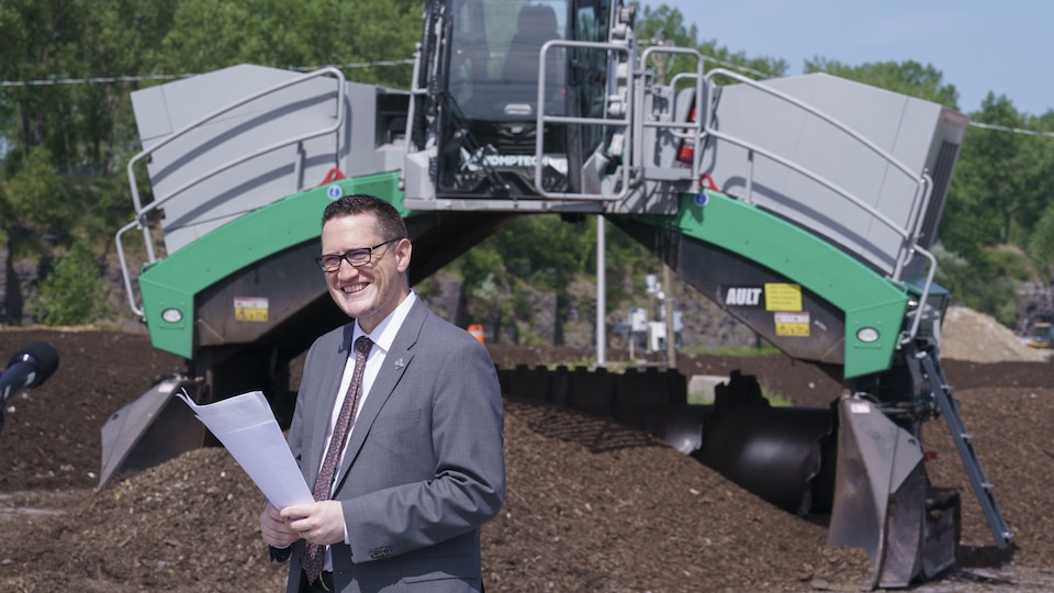 Un homme se tient debout en souriant devant de la machinerie destinée à l'aération du compost.