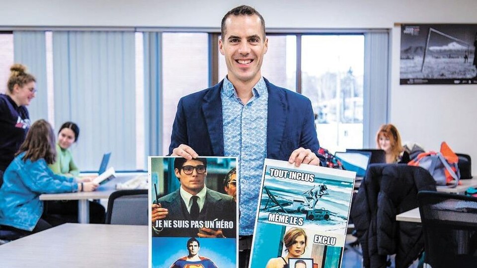 Benoît Dumais sourit à la caméra en tenant deux affiches sur lesquelles on peut y voir deux de ses «memes».