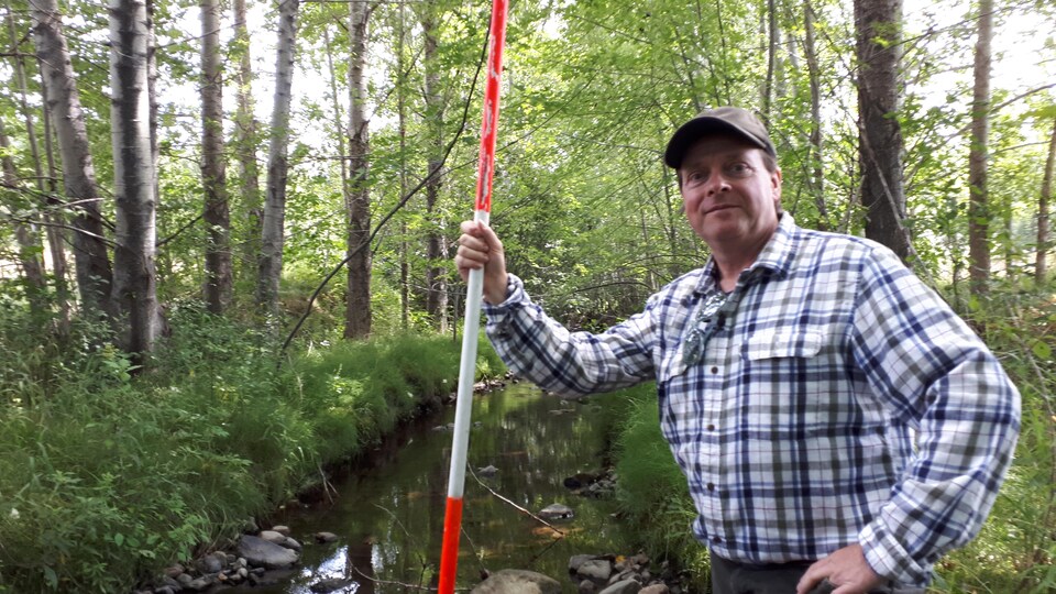Le chercheur tient une grande règle à la main au milieu d'un ruisseau entouré de peupliers.