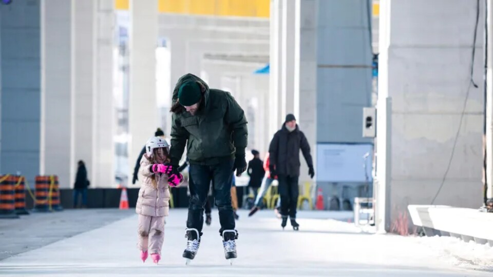 Un père patine en tenant la main de sa fille. Ils sont entouré des pylônes de béton qui soutiennent l'autoroute Gardiner.