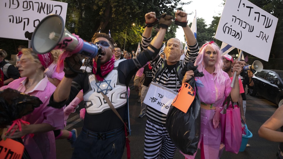Des manifestants entourent un sosie du premier ministre habillé en prisonnier et menotté.