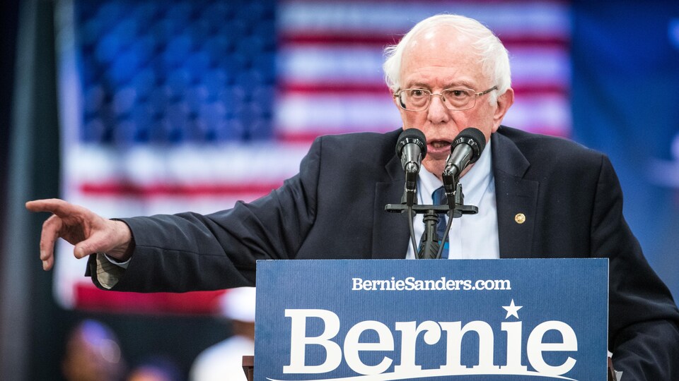 Un homme portant des lunettes parle derrière un lutrin en levant le bras droit. Une pancarte sur laquelle il est écrit «Bernie» est posée devant lui.