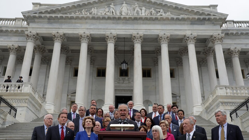 Les sénateurs démocrates entourent Chuck Schumer, qui parle au micro devant un lutrin, sur les marches du Capitole.