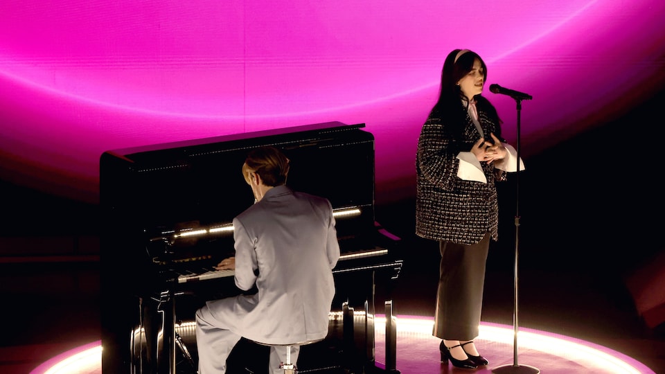 Un homme au piano et une femme au micro sont sur une scène circulaire illuminée en rose. 