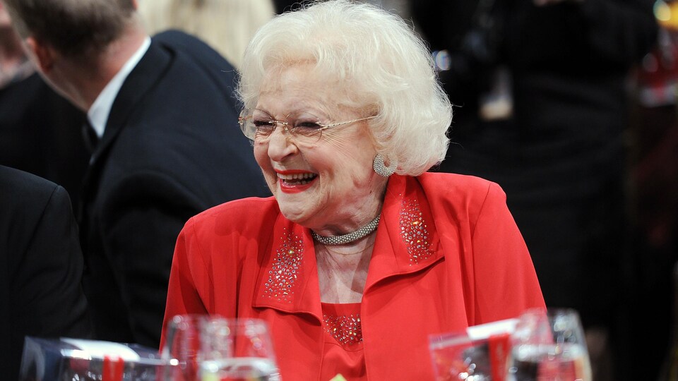 L'actrice Betty White, vêtue d'une tenue rouge, assise à une table lors d'un événement mondain.