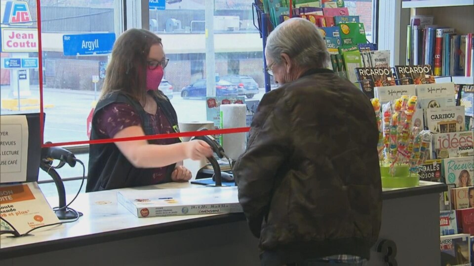 Une caissière, derrière une vitre de plexiglas, sert un client avec un masque.