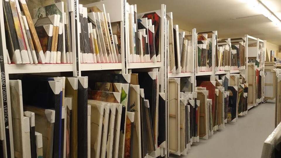Des peintures sont tablettées en série dans une grand bibliothèque sur des étagères. 
