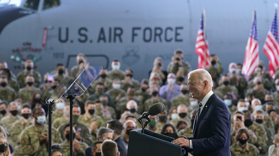 Joe Biden, souriant, devant un lutrin, avec un avion de la US Air Force et des drapeaux américains en arrière-plan, s'adresse à des militaires américains.