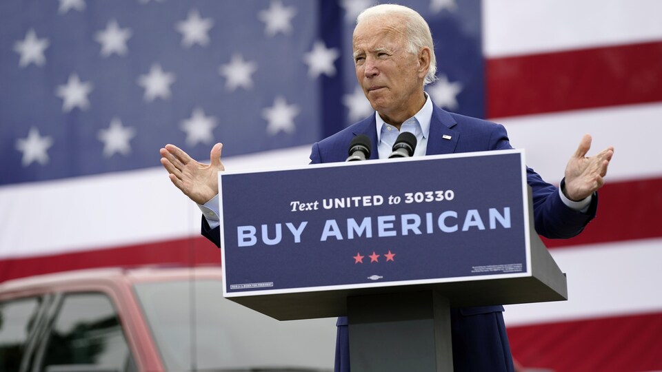 Joe Biden prononce un discours devant un lutrin sur lequel on peut lire « Buy American ». Derrière lui, un drapeau américain.