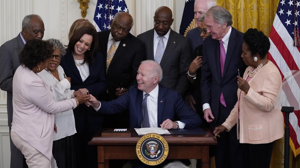 Au milieu d'un groupe d'élus réunis à la Maison-Blanche, une représentante tend un stylo à Joe Biden, qui sourit, tout comme Kamala Harris.