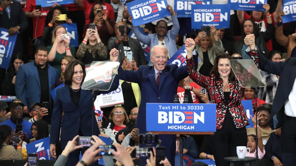 Entouré de Kamala Harris et de Gretchen Whitmer, Joe Biden lève les bras devant une foule de partisans, dont certains tiennent des affiches invitant à voter pour le candidat démocrate.