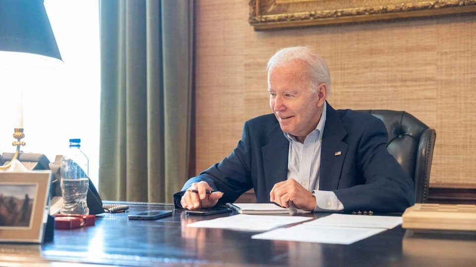 Joe Biden, vêtu d'un costume bleu, est assis à un bureau, stylo en main. 