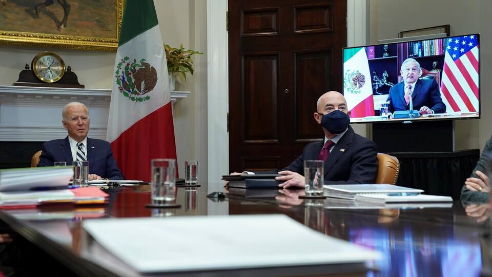 Le président Joe Biden, assis au bout de leur table devant un drapeau mexicain, avec Alejandro Mayorkas à ses côtés, et Andrés Manuel López Obrador, sur un écran, entre des drapeaux américain et mexicain.