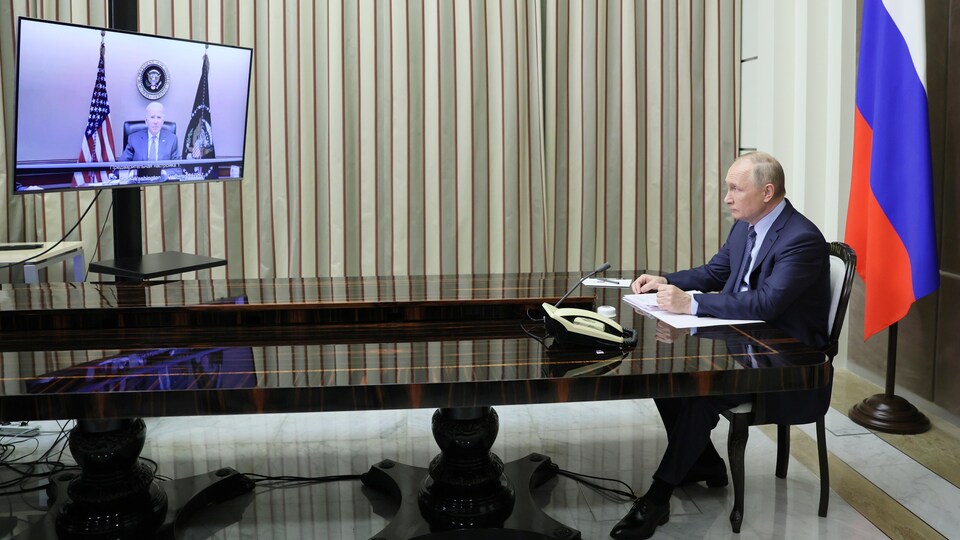 Le président russe, assis au bout d'une longue table, regarde un écran placé à distance et sur lequel on voit Joe Biden.