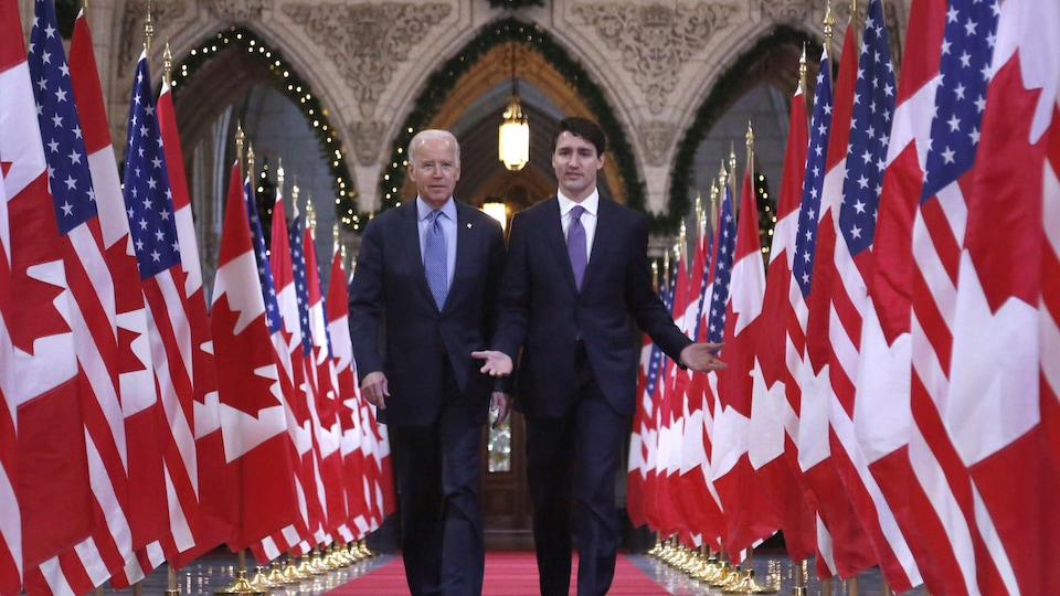 Joe Biden et Justin Trudeau marchent côte à côte au milieu d'une rangée de drapeaux canadiens et américains.