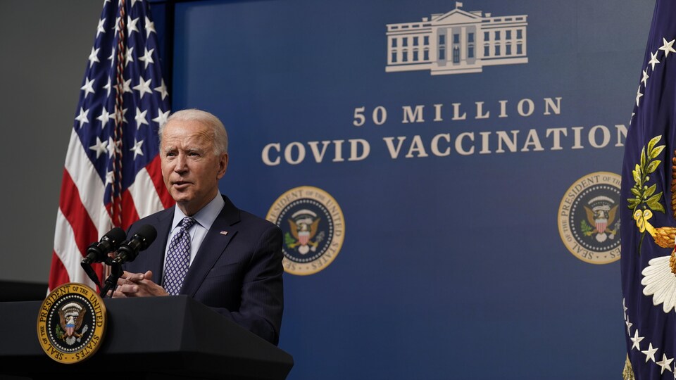 Joe Biden, au micro, s'adresse aux journalistes, devant un drapeau américain et un énorme panneau sur lequel on peut lire: «50 millions – Vaccination contre la COVID ».