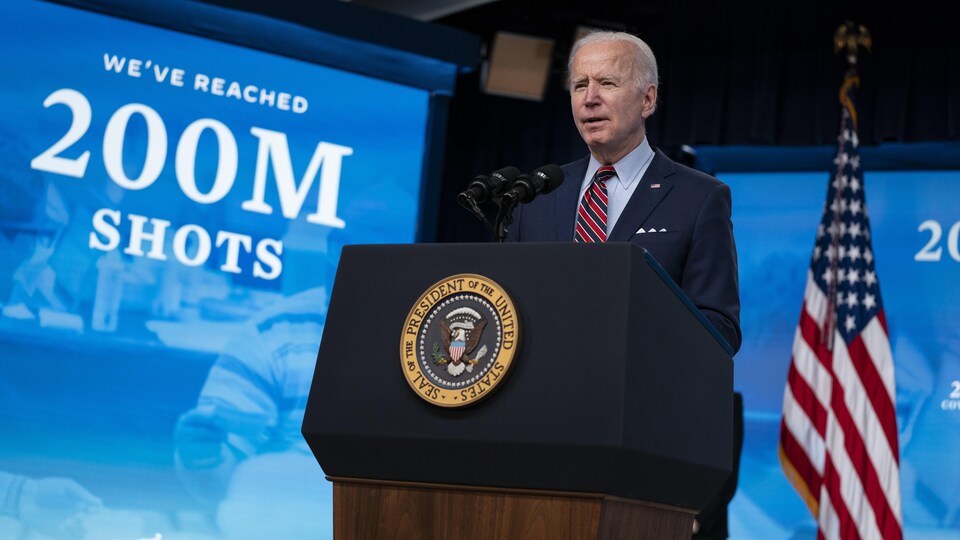 Joe Biden, regardant la caméra, à côté d'un drapeau américain, devant un large panneau disant : « Nous avons atteint 200 millions de doses. »