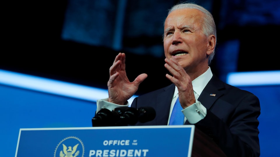 Les mains levées, le président désigné Joe Biden regarde la caméra devant un lutrin où est écrit « Bureau du président désigné ».