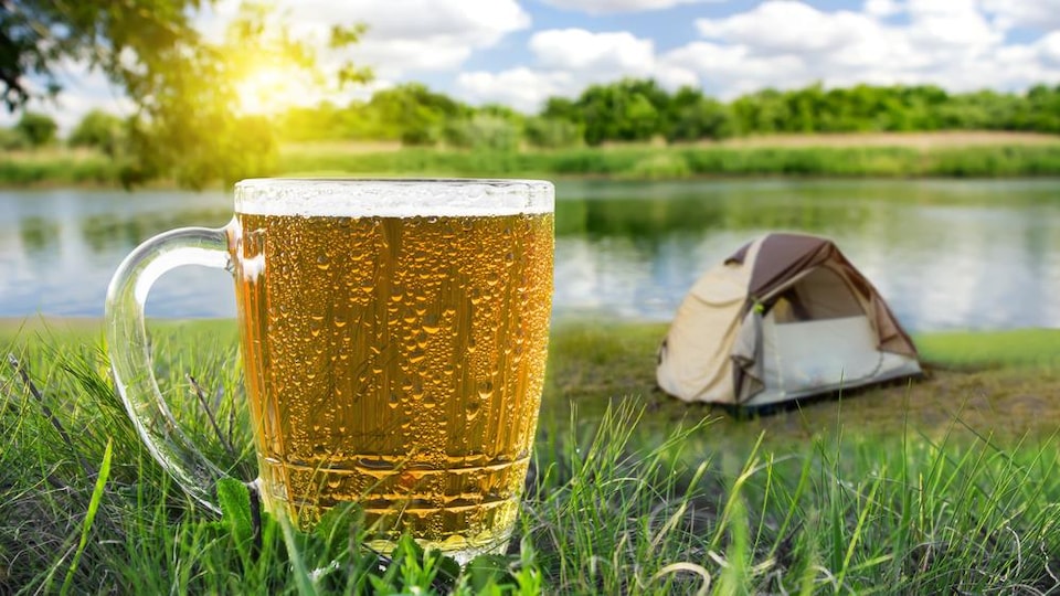 Un verre de bière froide en pleine nature avec, en arrière-plan, une tente, un lac ainsi qu'un soleil brillant