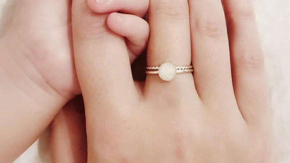 La main d'un bébé tient l'index de sa mère. Sur le majeur est enfilée une bague surmontée d'une perle de couleur laiteuse.