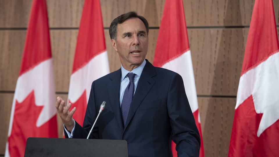 Bill Morneau gesticule en parlant derrière un lutrin et devant des drapeaux du Canada.