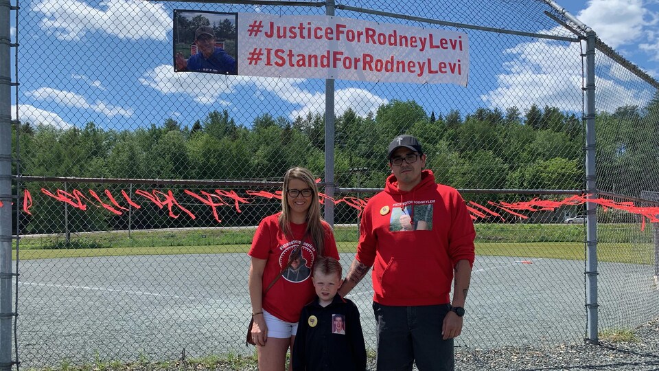 Becky Levi (la nièce de Rodney Levi), son fils et Bill Ward, le chef de la communauté de la Première nation Metepenagiag devant une clôture de terrain de baseball à l'extérieur.