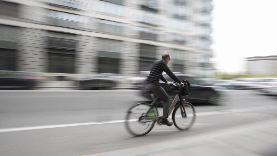 Un cycliste fend l'air, sur un Bixi, dans une rue de Montréal, tandis qu'une voiture passe à côté de lui.