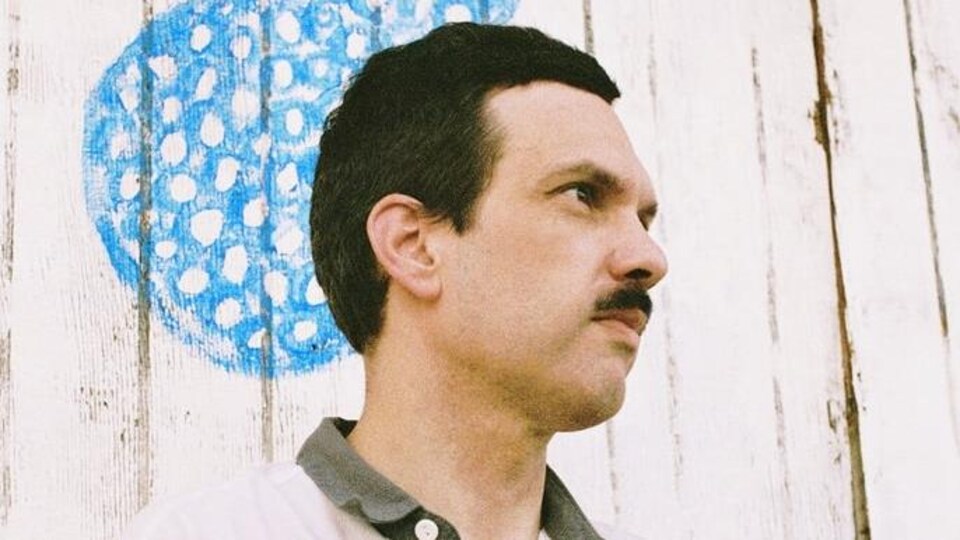 Un homme portant la moustache vue de biais, un mur peint derrière lui.