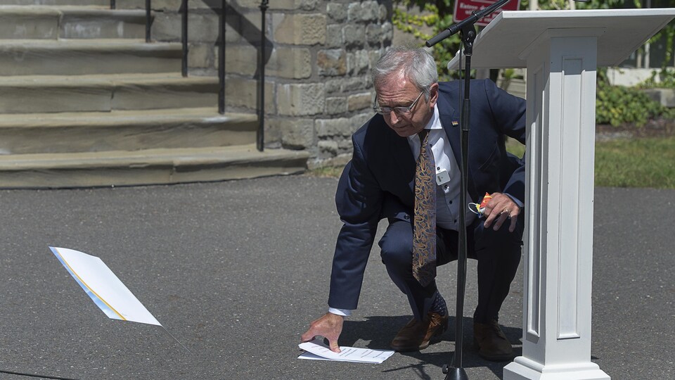 Blaine Higgs est agenouillé devant un lutrin et il ramasse par terre des feuilles de papier qui sont parties au vent.