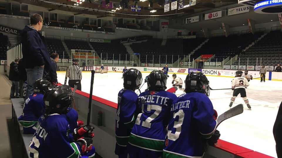 De jeunes hockeyeurs de deux équipes pendant un tournoi au Colisée de Rimouski, certains sont sur le banc des joueurs, d'autres sur la glace.